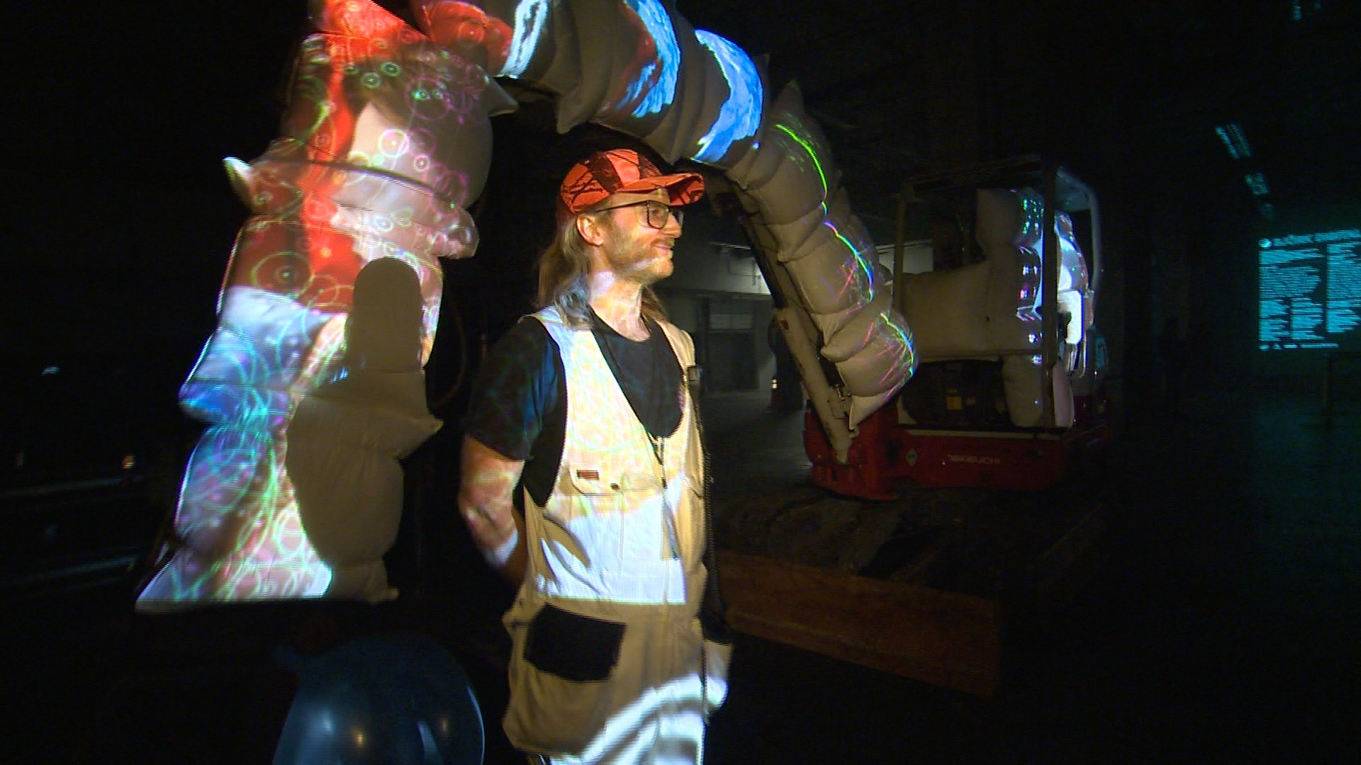 Alex Czetwertynski at night beside an illuminated inflatable installation and an excavator during Day for Night build-out