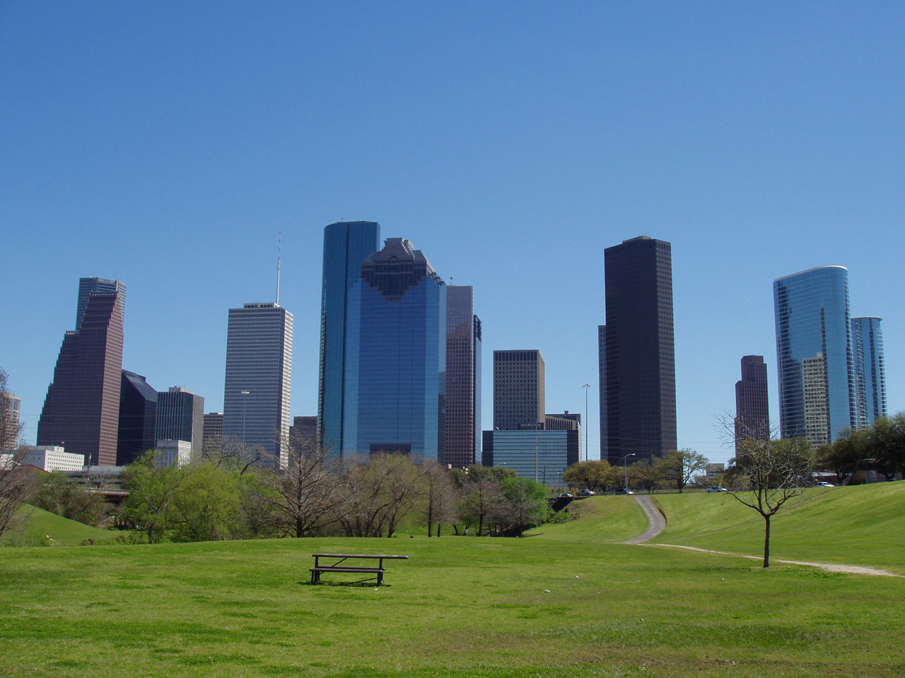 The downtown Houston skyline seen from the grassy slopes of Eleanor Tinsley Park under a clear blue sky, with a picnic table and a path across the lawn in the foreground.