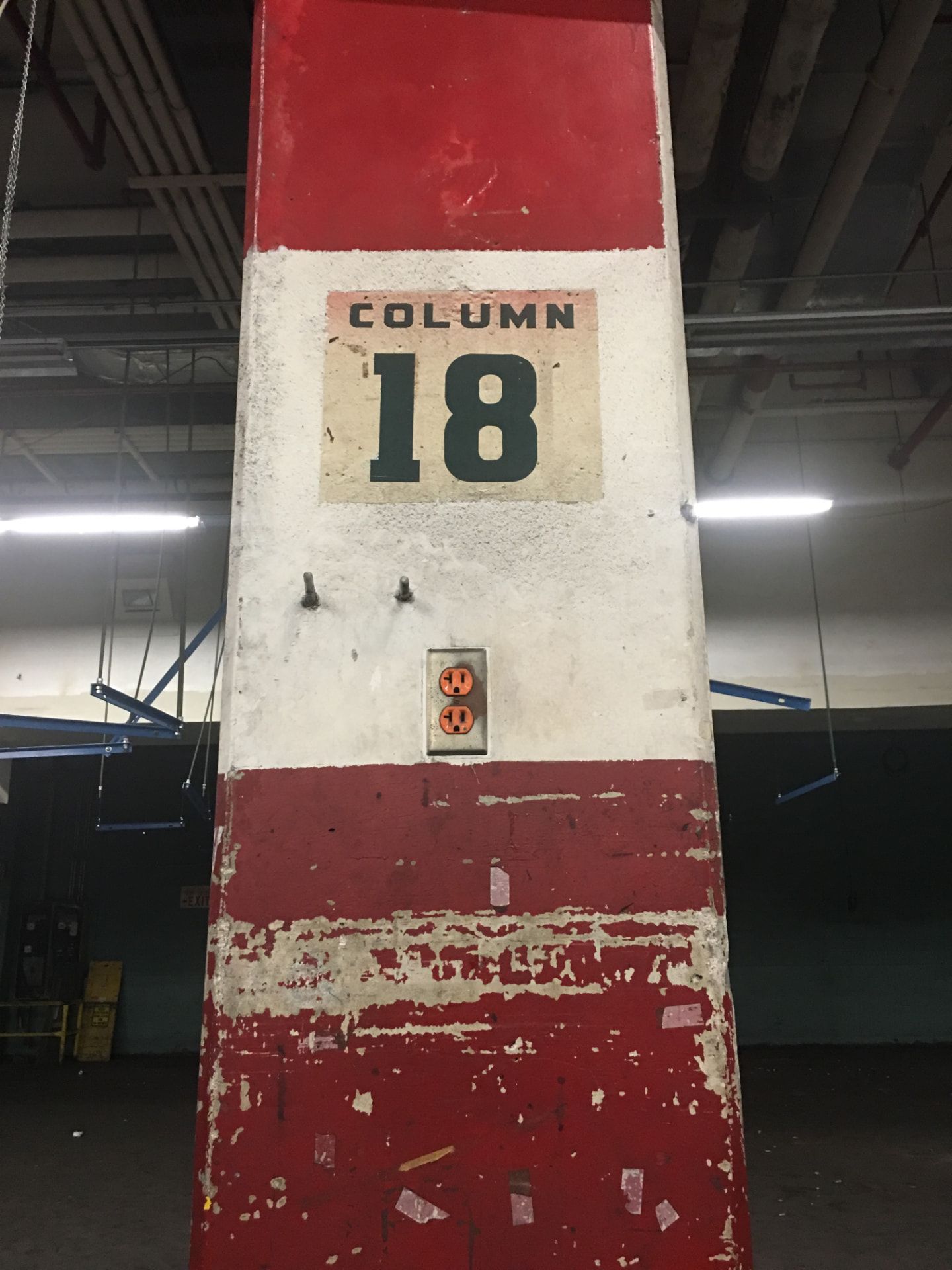 A stenciled COLUMN 18 marker on a red and white painted concrete column inside the former post office, with a worn electrical outlet below.