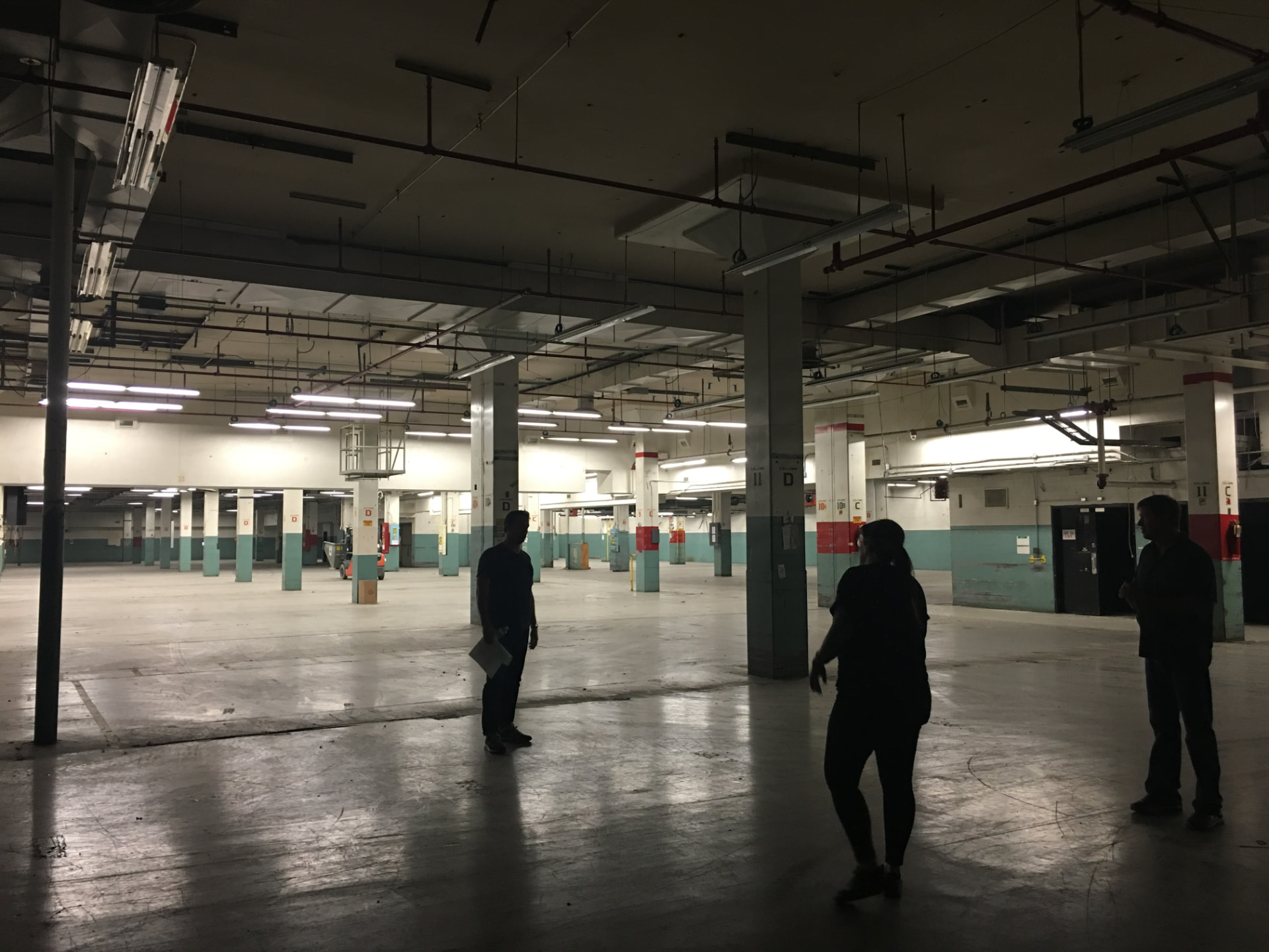 Three people in silhouette walking the dimly lit empty floor of the former post office beneath exposed ductwork and column rows.