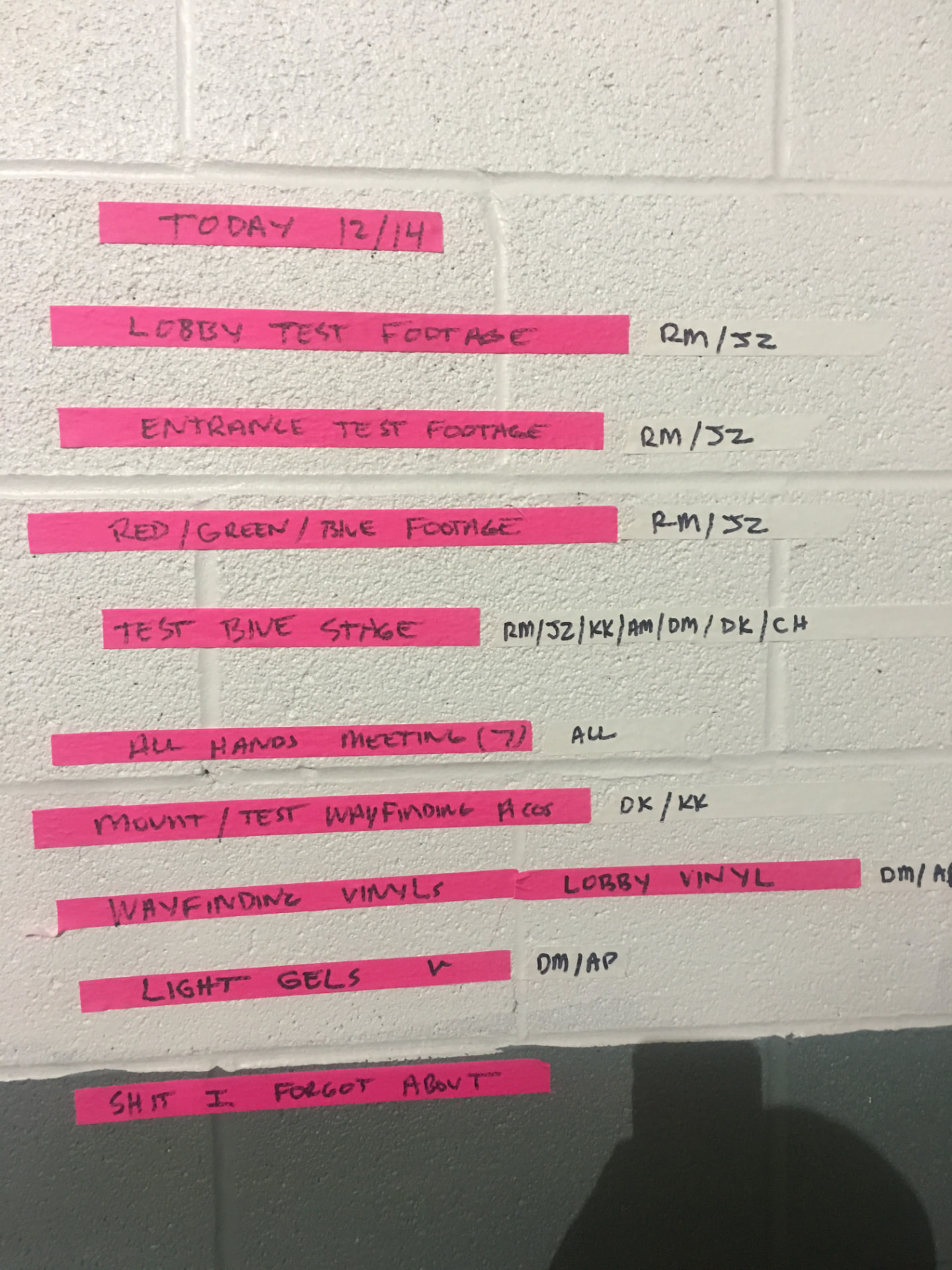 A handwritten production to-do list on pink tape strips stuck to a cinder-block wall, headed TODAY 12/14, with tasks including lobby test footage, entrance test footage, test Blue Stage, an all-hands meeting, wayfinding vinyls and light gels, with crew initials beside each task.