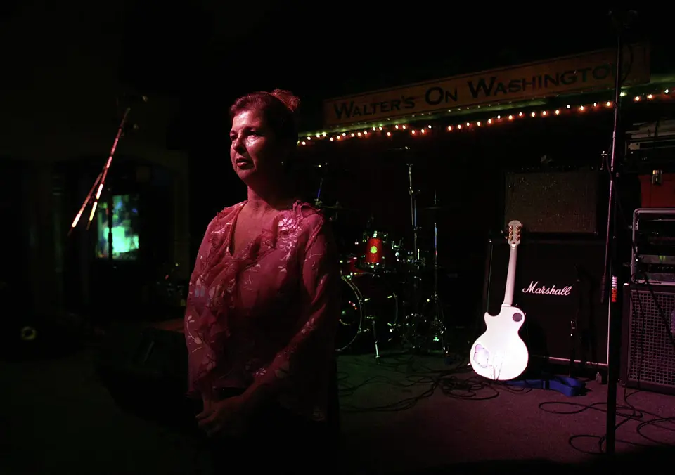 Dark stage with a woman standing beside a drum kit, a white guitar and a Marshall amplifier under a Walter's on Washington banner.
