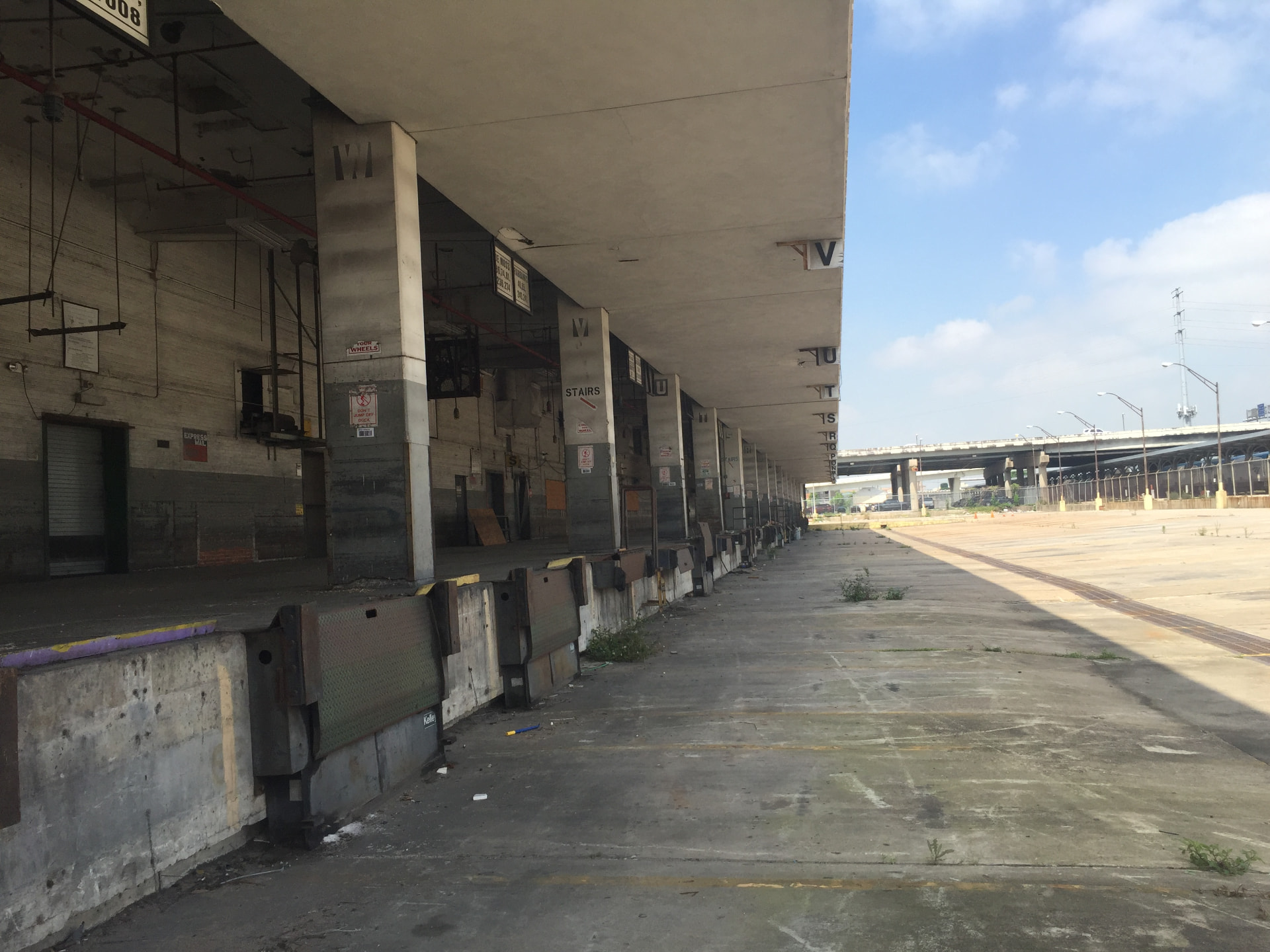 The long covered loading dock of the former post office with lettered bays and dock levelers, the elevated freeway visible beyond the empty lot.