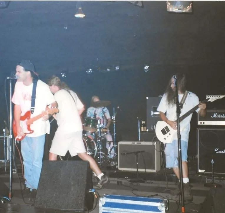 Stage view at Instant Karma: two guitarists in front, a drummer behind, Marshall amplifiers stacked to the right.