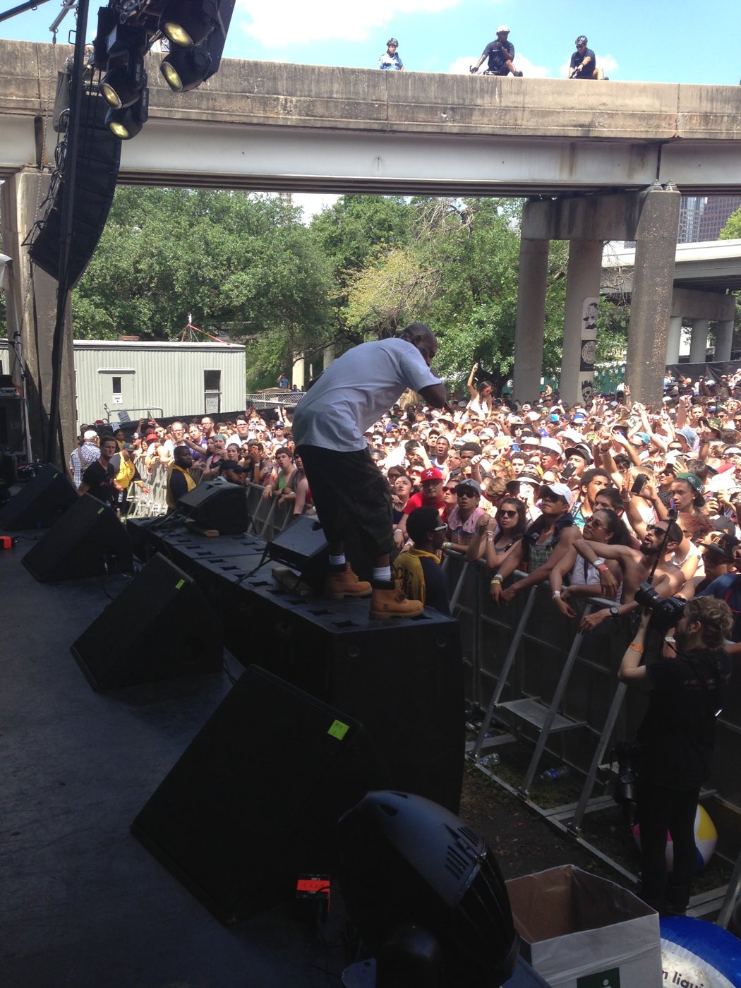 Performer on stage monitors above a packed daytime festival crowd beneath a Houston overpass.