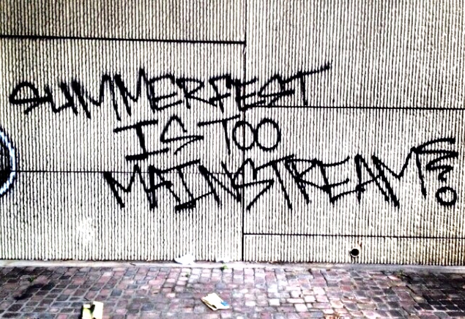 Black spray paint on a ribbed concrete wall above brick paving, reading “SUMMERFEST IS TOO MAINSTREAM?”. Photographer and date not established.