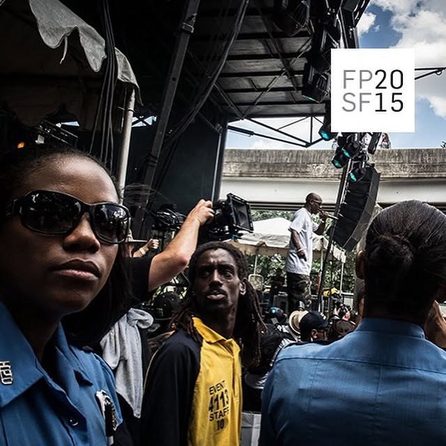 Stage-side crowd under a highway overpass: an officer in sunglasses at left, a person in a yellow EVENT STAFF shirt at centre, camera and lighting rigging overhead, with a printed FP20 / SF15 mark at upper right; date and photographer not established.