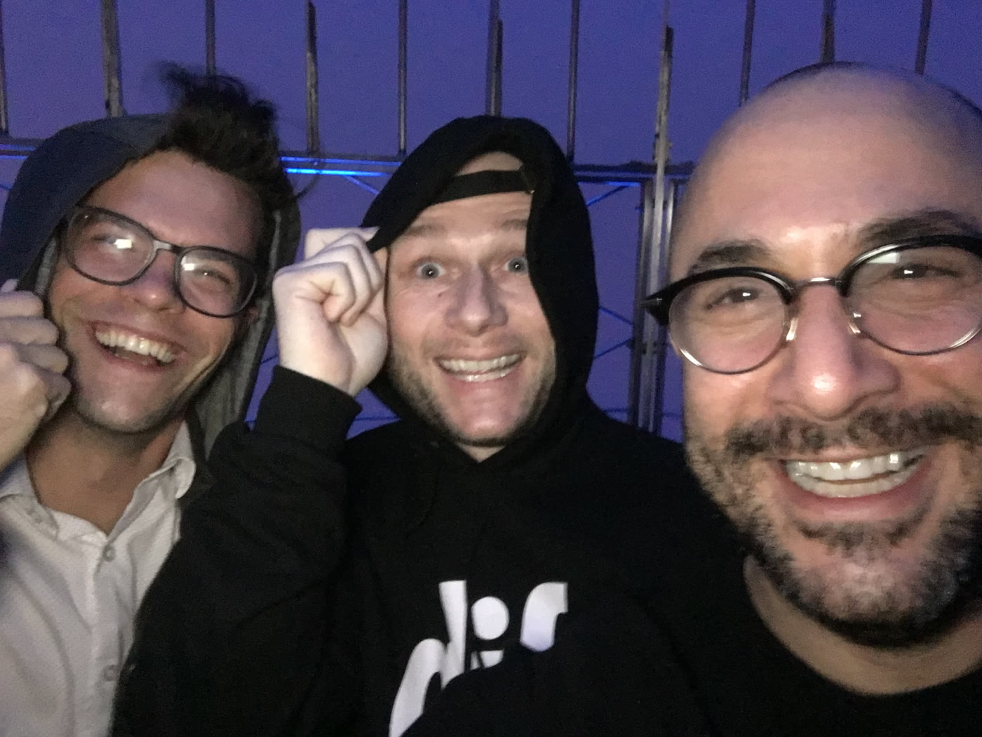Three people shoulder to shoulder at night, smiling at the camera, with purple-lit truss and rigging behind them; the person at left wears glasses and a light shirt, the person at centre wears a black hoodie and cap, the person at right wears dark-framed glasses; date, location and photographer not established.